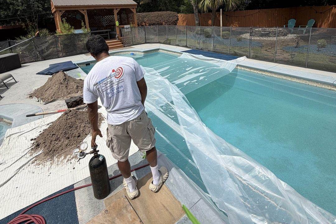 A technician at a poolside with a pressure-test cylinder and hose, the pool sheeted over, sand piles and plywood on the deck