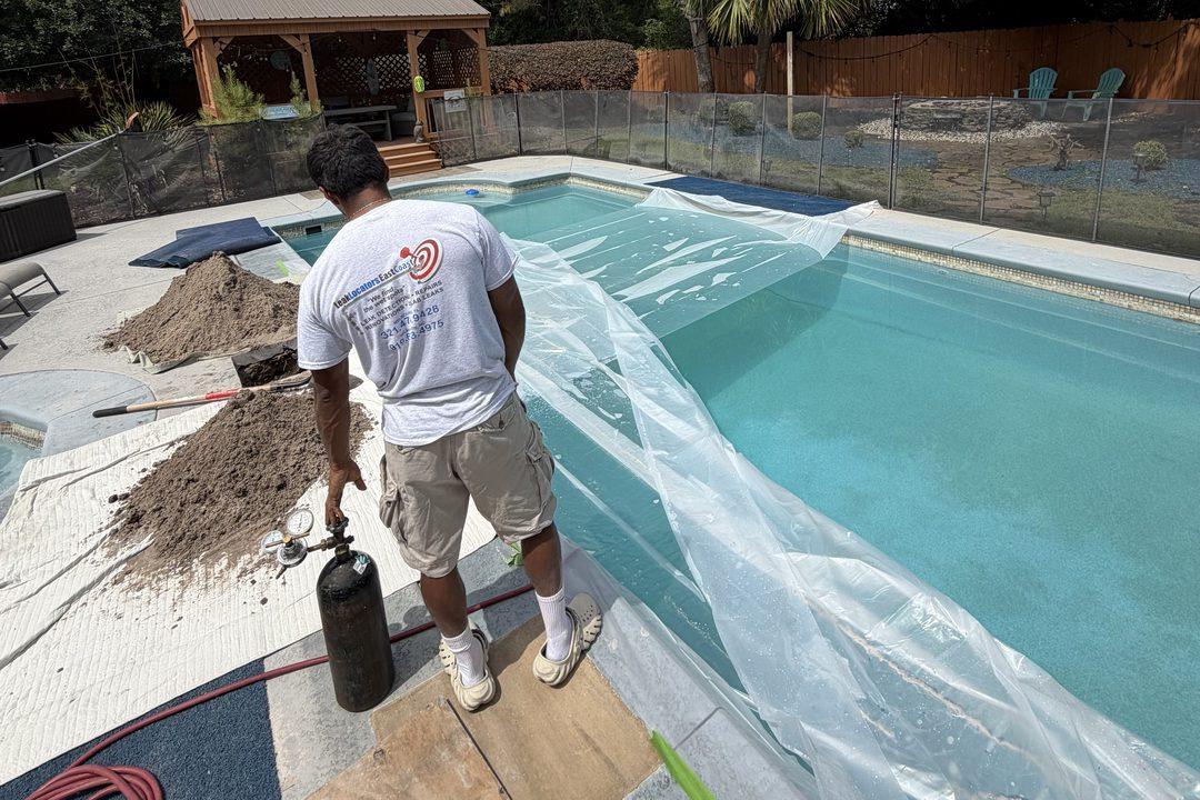 A technician at a poolside with a pressure-test cylinder and hose, the pool sheeted over, sand piles and plywood on the deck