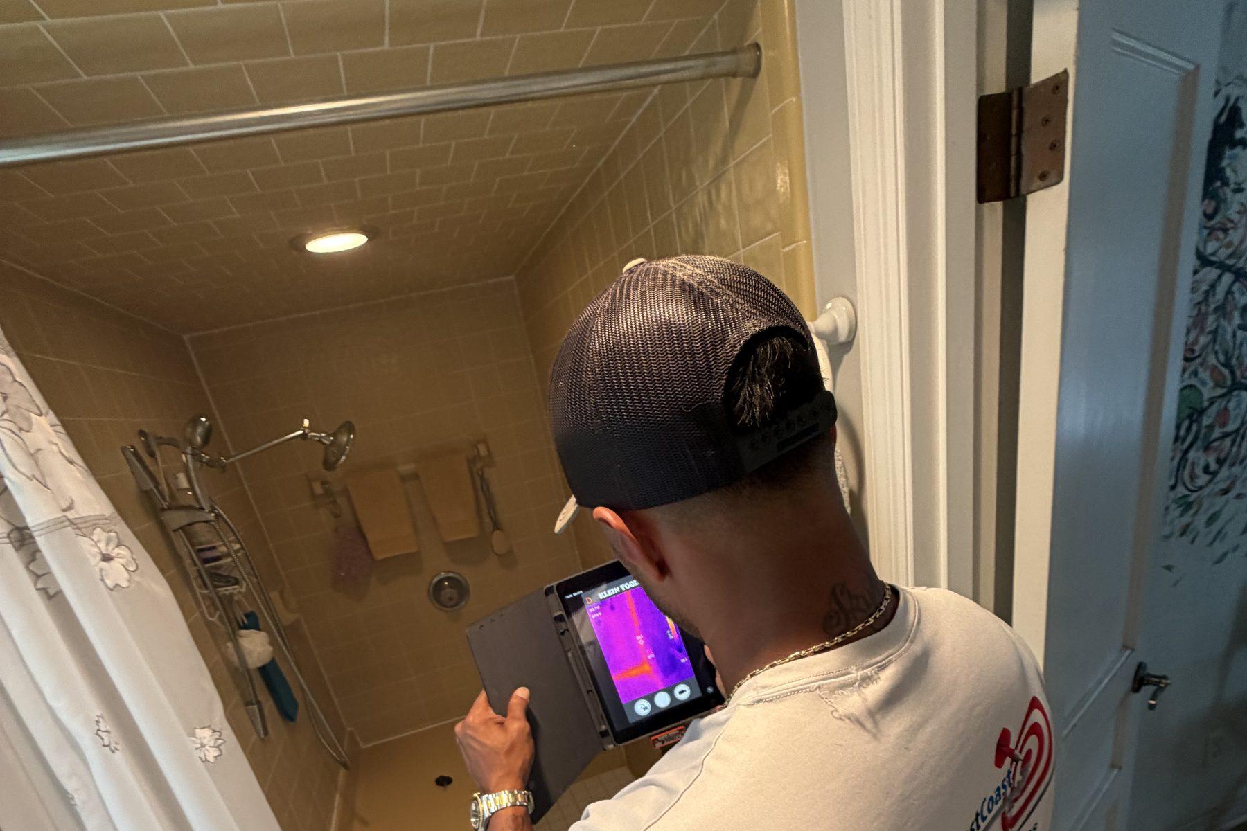 A technician holding a thermal imaging camera with the display visible, aimed at a tiled shower area